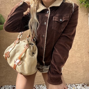 May include: A brown corduroy jacket with a cream shearling collar and trim. The jacket features button closures and two front pockets. A beige handbag with brown leather straps and silver hardware is also visible.
