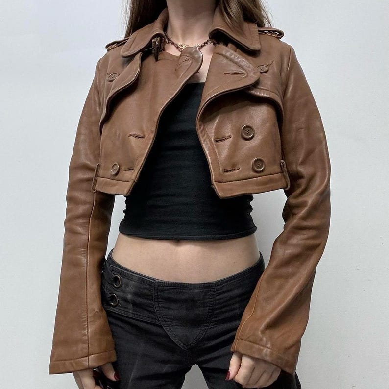 May include: A cropped brown leather jacket with a double-breasted design. The jacket features a notched collar, button closures, and long sleeves. The jacket is worn over a black top and paired with black pants.