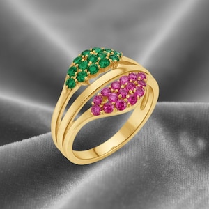 Könnte beinhalten: Ein goldener Ring mit zwei Bändern, die jeweils mit einem Cluster aus Edelsteinen besetzt sind. Ein Cluster zeigt leuchtend grüne Steine, der andere leuchtend rosa Steine. Der Ring hat eine polierte Oberfläche.