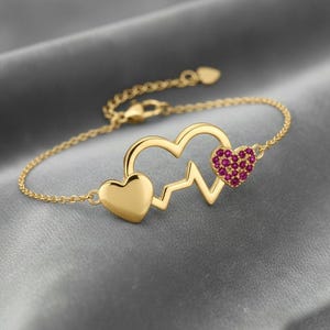 Könnte beinhalten: Goldfarbenes Armband mit Herzmotiv. Das Armband hat eine Kette mit Verschluss und einen herzförmigen Anhänger. Ein Herz ist massiv, das andere mit kleinen, roten Edelsteinen gefüllt. Das Design beinhaltet eine Herzschlaglinie.