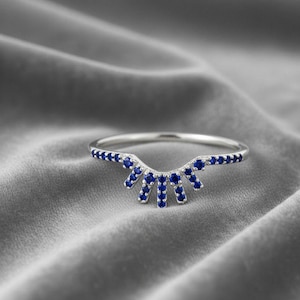 Könnte beinhalten: Ein silberfarbener Ring mit geschwungenem Design, verziert mit zahlreichen kleinen, leuchtend blauen Edelsteinen. Der Ring hat ein schmales Band, und die Edelsteine sind in einem sonnenförmigen oder fächerartigen Muster angeordnet. Der Ring liegt auf einem weichen, grauen Stoff.