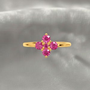 Könnte beinhalten: Ein goldener Ring mit einem floralen Design aus fünf leuchtend rosa Edelsteinen. Der Ring hat ein dünnes, poliertes Goldband, und die Edelsteine sind in einem kreuzartigen Muster angeordnet. Der Ring wird vor einem grauen Hintergrund präsentiert.