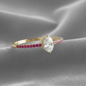 Peut inclure: Une bague en or avec un diamant de taille ovale en pierre centrale. La bande est ornée de petites pierres précieuses rouges rondes. La bague est présentée sur un fond gris, soulignant son design élégant.