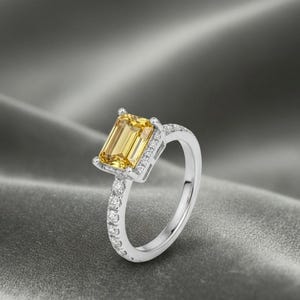 Könnte beinhalten: Ein silberner Ring mit einem großen, rechteckigen gelben Edelstein. Das Band ist mit kleinen, klaren Edelsteinen besetzt. Der Ring ist vor einem grauen, strukturierten Hintergrund platziert.