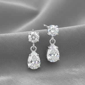 May include: A pair of silver drop earrings featuring clear, sparkling round and pear-cut gemstones. The earrings are presented on a grey, textured surface. The design is classic and elegant.