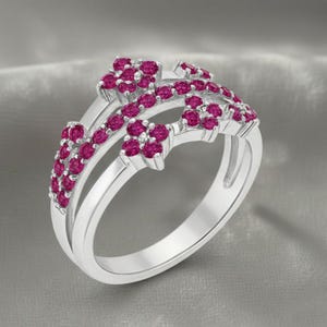 Könnte beinhalten: Ein silberner Ring mit floralem Design, besetzt mit zahlreichen kleinen, runden, magentafarbenen Edelsteinen. Der Ring hat einen geteilten Schaft und eine zarte, aufwendige Fassung, die ein funkelndes und elegantes Aussehen erzeugt.