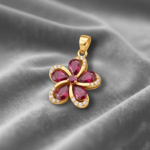 May include: A gold pendant shaped like a flower with five petals. Each petal contains a teardrop-shaped, deep red gemstone, edged with small, clear crystals. The flower's centre also has a smaller red gemstone.