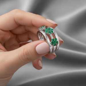 Könnte beinhalten: Ein silberner Ring mit drei Bändern, verziert mit Clustern aus kleinen, leuchtend grünen Edelsteinen. Der Ring hat ein Blumenmuster, wobei die Edelsteine so angeordnet sind, dass sie Blumen ähneln. Der Ring wird von einer Person gehalten.