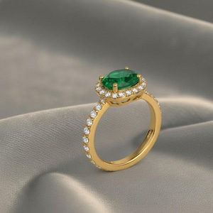 Könnte beinhalten: Ein goldener Ring mit einem ovalen Smaragd, umgeben von einem Kranz aus kleinen Diamanten. Der Ring hat ein mit Diamanten besetztes Band. Das Design ist elegant und luxuriös. Der Ring wird vor einem weichen, grauen Hintergrund präsentiert.