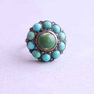Può includere: Anello d'argento con una pietra verde centrale circondata da pietre turchesi. L'anello ha un design vintage, con un'incastonatura dettagliata. Lo sfondo è chiaro.