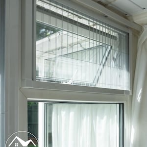 May include: A white window frame featuring a retractable pleated insect screen installed in the upper section. The screen has a grid pattern and is mounted within a white casing. A circular logo with the text HILT PRO IDEAL LIFE is visible.
