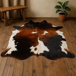 May include: A brown, black, and white cowhide rug with a natural, irregular shape. The rug is laid out on a wooden floor.