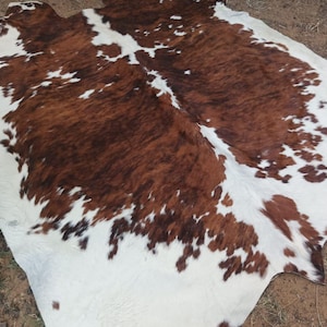 May include: A brown, black, and white cowhide rug with a natural, irregular shape. The rug is laid out on a wooden floor.