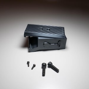 Puede incluir: Un dispositivo rectangular negro con un patrón de fibra de carbono en la parte superior, posiblemente un accesorio de cámara. Varios tornillos negros de diferentes tamaños están dispuestos debajo del dispositivo.
