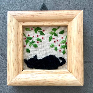 以下が含まれることがあります： 赤い実のついた緑の葉の枝の下で眠る黒い猫の刺繍が、明るい木製の額縁に入っています。刺繍は白いリネン生地に施されています。