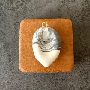 May include: A teardrop-shaped pendant featuring a marbled pattern of white and dark gray tones. The surface has a smooth, matte texture with small pinhole details. A small, circular gold-toned metal jump ring is attached to the top center.