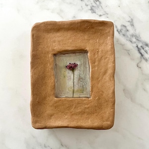 以下が含まれることがあります： 手作りの長方形のセラミック作品で、四角い窪みがあります。四角には、茎の上の単一の乾燥した赤い花の絵が描かれています。フレームは暖かく土のような茶色で、背景は明るくニュートラルな色調です。