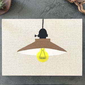 以下が含まれることがあります： 吊り下げ式のランプの様式化されたイラストが描かれたキャンバスアート作品。 ランプは茶色のシェード、黒色の器具、黄色の電球を備えています。 背景はニュートラルなベージュ色です。
