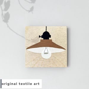 以下が含まれることがあります： 黒いコードと黒い金属製のベースが付いた茶色の照明器具が、白い電球と共に描かれた茶色のテキスタイルアート作品です。