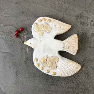 以下が含まれることがあります： 白いセラミックの鳩の置物。金色のアクセントと赤い実の枝がくちばしにあります。鳥は質感のある翼と体のディテールを持ち、灰色の背景に配置されています。