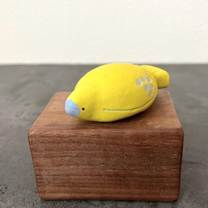 May include: A handcrafted, yellow ceramic bird figurine with a blue beak and small gray accents, resting on a small, square, wooden block. The bird has a simple, whimsical design, and the block has a natural wood grain.