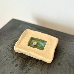 以下が含まれることがあります： 手作りの正方形のセラミック皿で、中央がくぼんでいます。 皿は薄いタン色で、長方形のインセットには、緑と青の背景に小さな白い花が描かれています。 この皿は、小さなアイテムを入れるために使用される可能性があります。