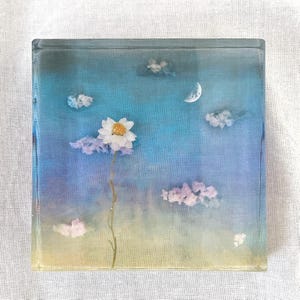 May include: Square resin art piece featuring a white daisy with a yellow center, set against a blue and yellow sky with fluffy cloud accents and a crescent moon. The artwork is encased in clear resin.