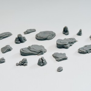 May include: A collection of 14 grey 3D printed rock formations of various sizes and shapes. The rocks are scattered on a white surface.