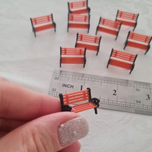 Könnte beinhalten: Miniatur-Parkbänke in Orange und Schwarz. Die Bänke haben ein Sitzdesign mit Lamellen und schwarze Armlehnen. Ein Lineal zeigt, dass die Bänke etwa 2,5 cm lang sind. Mehrere Bänke sind auf einer weißen Oberfläche verstreut.