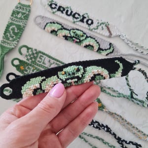 May include: Handmade friendship bracelets in various designs. One bracelet is black with a green snake design. Other bracelets are green and white with the word "CRUCIO" and "SLY" on them. The bracelets are made of woven thread.