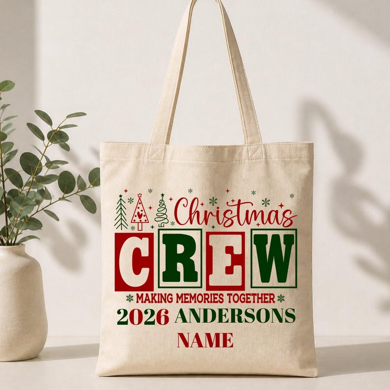 May include: A beige canvas tote bag featuring a festive graphic with the red and green text "Christmas CREW" above "MAKING MEMORIES TOGETHER", "2026 ANDERSONS", and "NAME", accented by small green and red Christmas tree and snowflake icons.