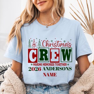 May include: A light blue t-shirt featuring a graphic with the red and green text "Christmas CREW" above "MAKING MEMORIES TOGETHER", "2026 ANDERSONS", and "NAME". The design includes small green and red Christmas tree icons and snowflakes.