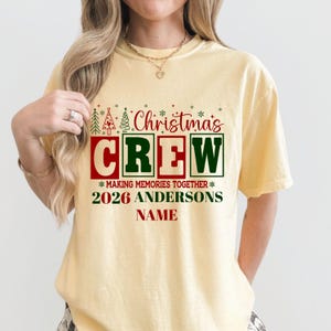 May include: A pale yellow short-sleeve t-shirt featuring a red and green graphic that reads Christmas CREW MAKING MEMORIES TOGETHER 2026 ANDERSONS NAME, decorated with small green and red Christmas tree and snowflake icons.