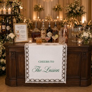 May include: A white fabric table runner with intricate lace trim edges, featuring the dark green printed text 'CHEERS TO The Lussiers'. The runner is draped over a dark wooden bar counter, which is set with various glass liquor bottles, cocktail shakers, and a framed signature cocktail menu sign.