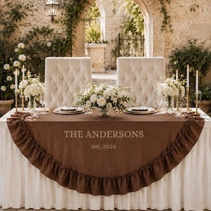 May include: A rectangular table draped in a white cloth with a brown, semi-circular ruffled runner featuring the text THE ANDERSONS est. 2026. The table is set with two tufted white chairs, white floral centerpieces, and tall gold candlesticks.