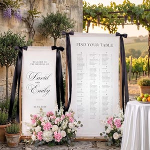 May include: Wedding signage with the text "Welcome to the Wedding of David and Emily" and "Find Your Table". The signs are framed in wood, adorned with black ribbons, and surrounded by floral arrangements of pink and white flowers.