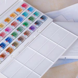 May include: A white watercolor paint set with an open lid revealing rows of colorful paint cakes. A separate palette and two paint brushes are also visible, suggesting a complete art kit for painting and drawing.
