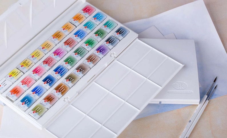 May include: A white watercolor paint set with an open lid revealing rows of colorful paint cakes. A separate palette and two paint brushes are also visible, suggesting a complete art kit for painting and drawing.