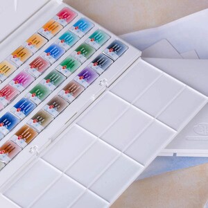 May include: A white watercolor paint set with an open lid revealing rows of colorful paint cakes. A separate white palette and two paint brushes are also visible. The paint cakes are arranged in a grid, showcasing a variety of colors.
