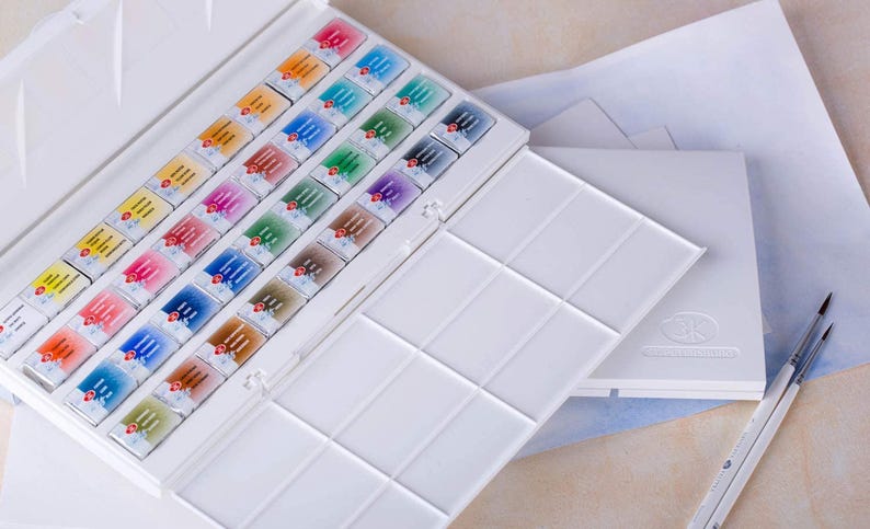May include: A white watercolor paint set with an open lid revealing rows of colorful paint cakes. A separate white palette and two paint brushes are also visible. The paint cakes are arranged in a grid, showcasing a variety of colors.