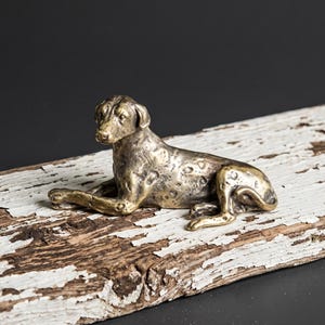 May include: A bronze-colored dog figurine in a resting pose. The dog is lying down with its front paws extended. The figurine has a textured surface and is placed on a weathered, white-painted wooden plank.