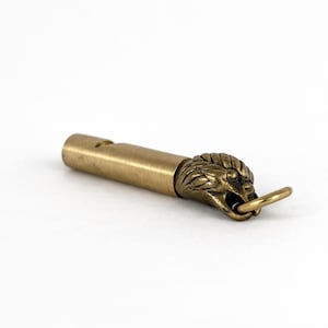 May include: A brass whistle featuring a lion's head. The whistle is cylindrical with a small ring attached to the lion's mouth. The lion's head is intricately detailed, and the whistle has a vintage aesthetic.