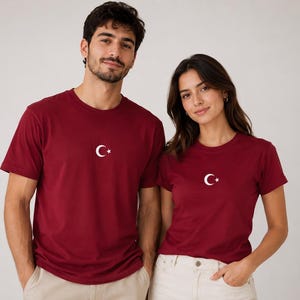 Könnte beinhalten: Ein dunkelrotes Kurzarm-T-Shirt mit einem kleinen weißen Halbmond und Stern-Symbol, das mittig auf der Brust platziert ist. Das Kleidungsstück verfügt über einen Rundhalsausschnitt und ein schlichtes, einfarbiges Design für einen lässigen Look.