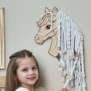 May include: A wooden horse head wall hanging with a macrame mane and decorative bows. The horse head is light brown with a white halter. A young person in a white dress with red and green floral embroidery is in the foreground.