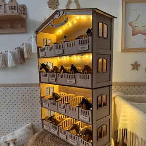 May include: A three-story wooden horse stable with miniature horses in stalls. The stable has a pitched roof and small windows. The stalls are labeled with names. The interior is illuminated with small lights. The stable sits on a wooden stool.