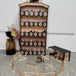 May include: A tall wooden toy horse stable with five levels of stalls, each featuring dark brown X-patterned doors. The structure is labeled 'Molly's Kinsley Ranch' and includes integrated lighting. A separate paddock area with wooden fencing, toy horses, and a small open-air shelter are arranged in front.