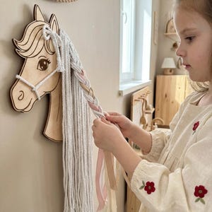 May include: Wooden horse head wall decor with a braided mane of white yarn and ribbons. The horse head is light brown with a detailed design. A child is braiding the yarn and ribbons. The wall is a light beige color.