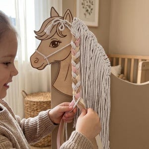 May include: A wooden horse head with a braided mane and a decorative design. The horse head is light brown with a white rope bridle and a long, flowing mane made of white yarn. The braid includes pink and tan ribbons.