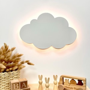 Op de afbeelding: Een witte, wolkvormige wandlamp met een warme gloed. De kinderkamerdecoratie omvat ook houten blokken met de tekst "BABY", drie houten konijnenfiguren en een houten "Animals Bus" speelgoed.