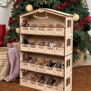 Puede incluir: Un establo de caballos de madera con múltiples compartimentos, cada uno con una figura de caballo en miniatura. El establo tiene un acabado marrón claro y está decorado con los nombres de los caballos. En el fondo se ve un árbol de Navidad decorado.